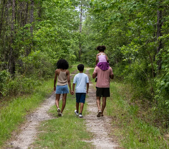 Family walking away from the camera on a wooded nature trail at Tributary, with a child riding on a parent's shoulders.