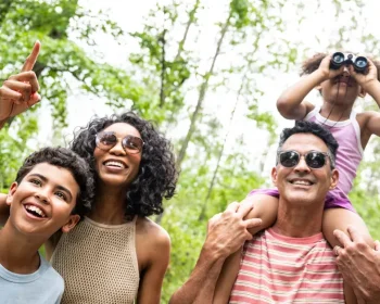 A joyful family explores a lush green park, with a child using binoculars from dad's shoulders while mom and son point at discoveries above.