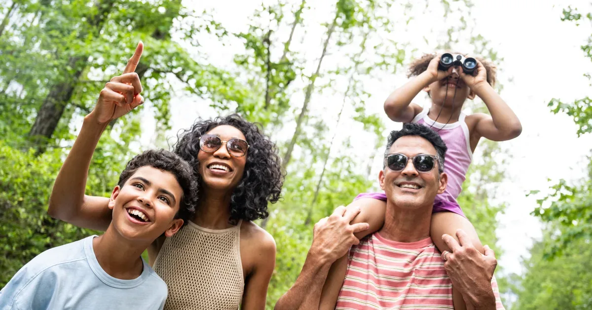 A joyful family explores a lush green park, with a child using binoculars from dad's shoulders while mom and son point at discoveries above.